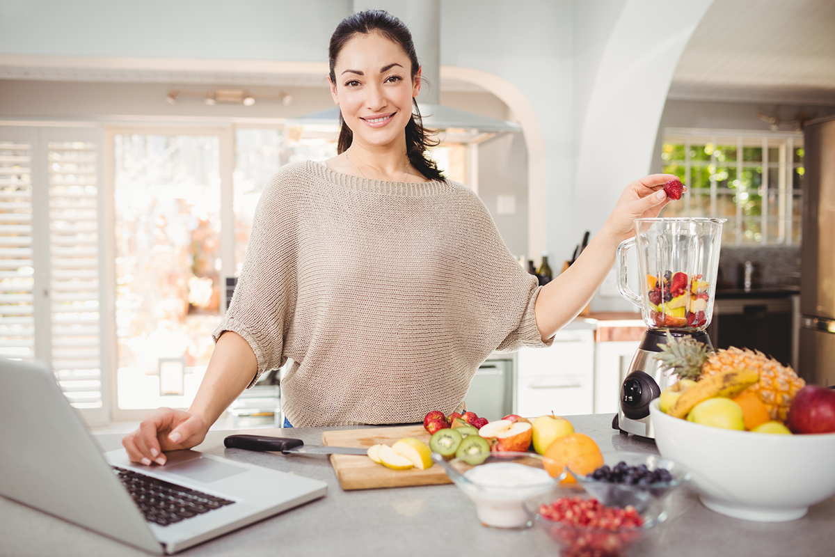 Portrait of smiling woman preparing fruit juice while working on laptop at table